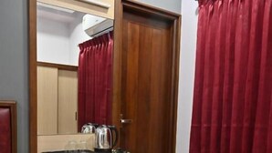 Ein kleines Hotelzimmer mit einer Holztür, einem kleinen Nachttisch, einem Spiegel, einer Ablagefläche mit einem Wasserkocher und roten Vorhängen.