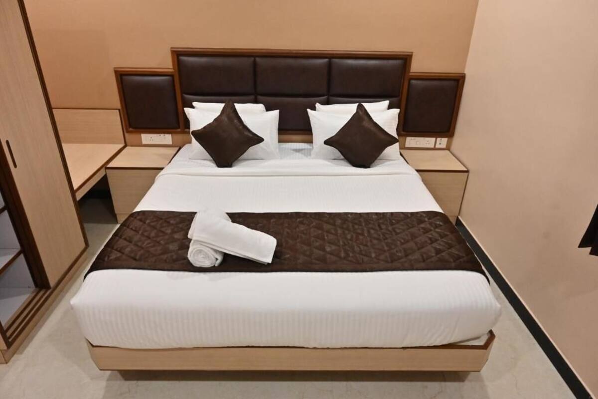 Una cama bien hecha con colcha marrón, sábanas blancas y dos almohadas marrones. Hay dos mesitas de noche con lámparas, y un armario de madera a la izquierda.