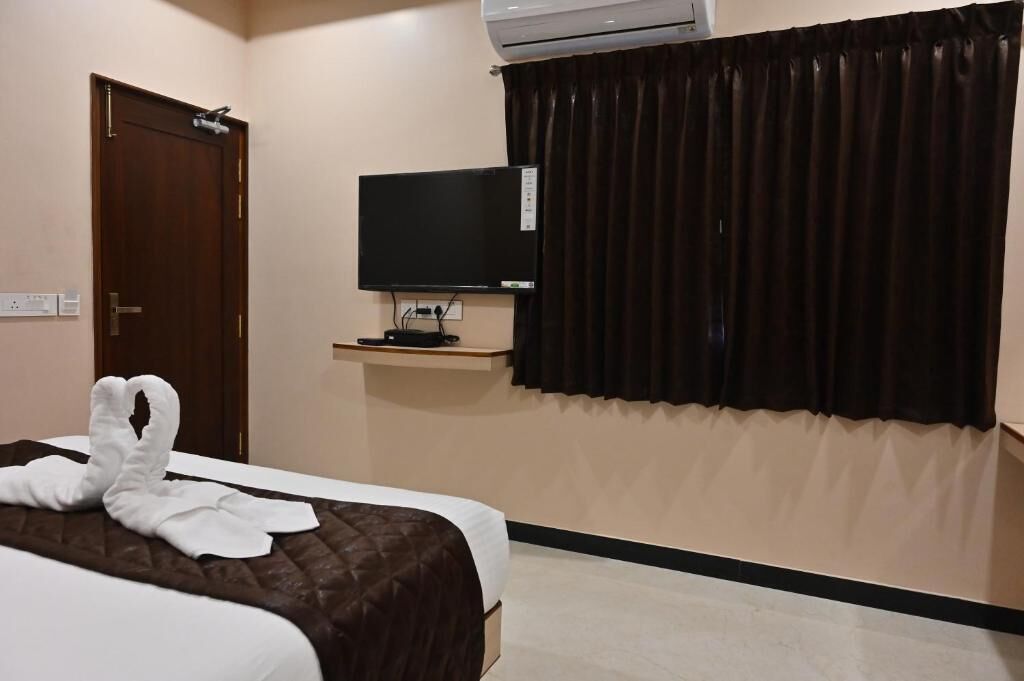 A hotel room with a bed, a flat-screen TV on a stand, a door, and a window with dark curtains.