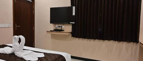 A hotel room with a bed, a flat-screen TV on a stand, a door, and a window with dark curtains.