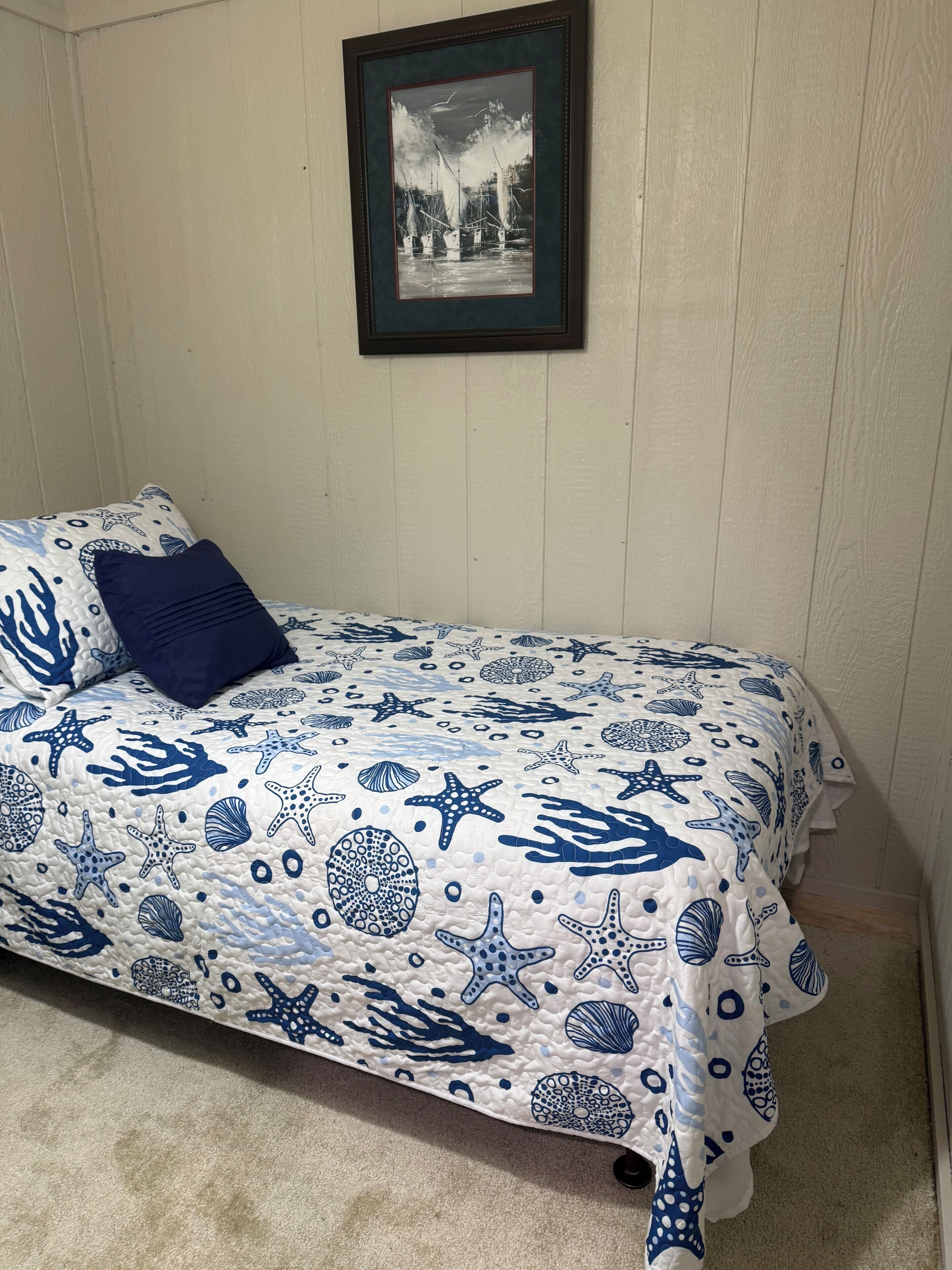 Een slaapkamer met een bed, voorzien van een beddegoed met een nautisch thema, een ingelijst plaatje aan de muur en één donker kussen.