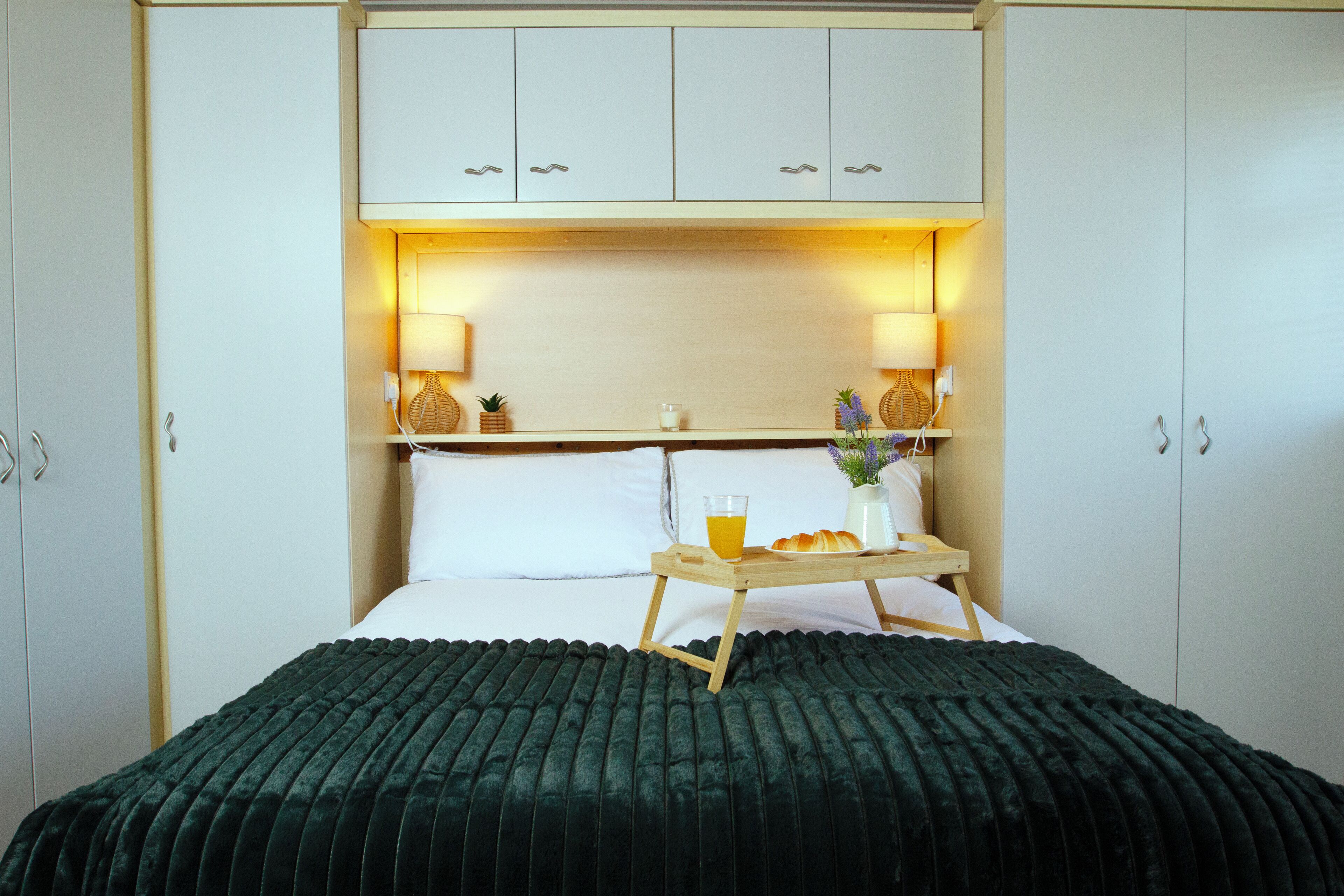 Una cama bien hecha con un edredón verde oscuro, una mesita de noche con una bandeja que sostiene un vaso de jugo de naranja y croissants, y dos lámparas fijadas a la pared.