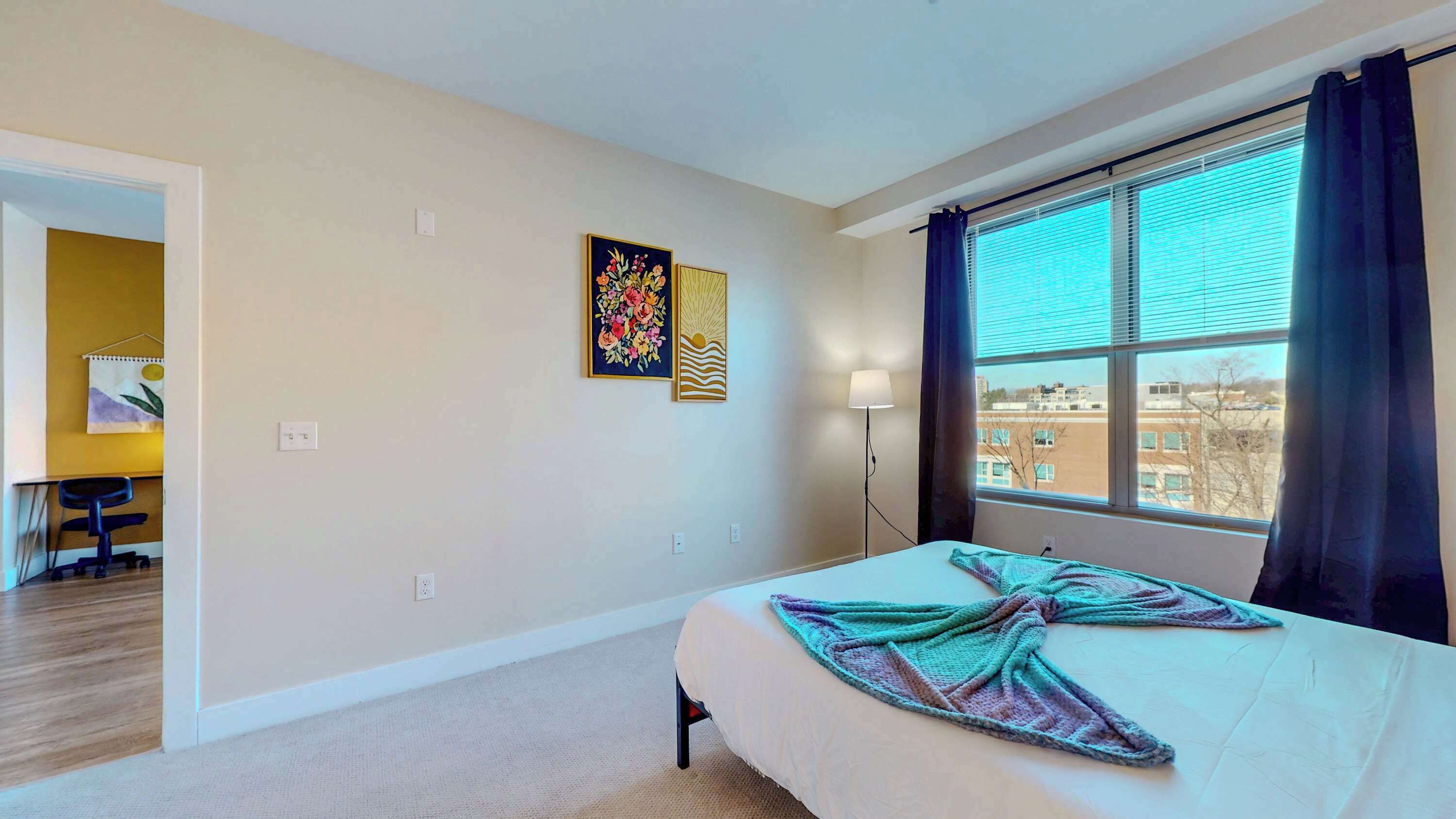 Habitación de hotel con cama, escritorio, silla, ventana con cortinas y dos obras de arte en la pared.