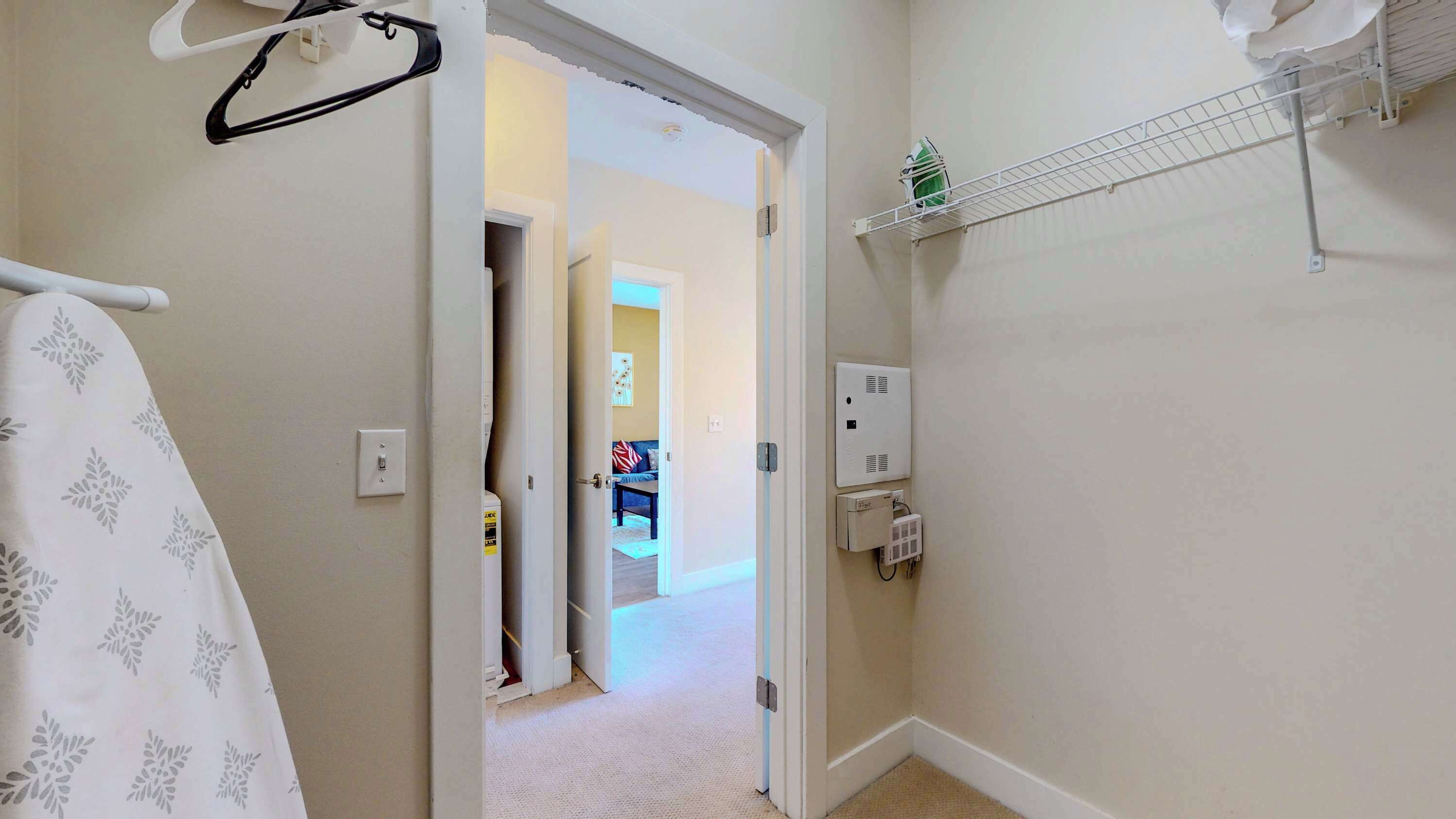 Un couloir avec une porte blanche donnant sur un salon, un porte-serviettes blanc, une étagère blanche avec une petite plante et une prise électrique fixée au mur.