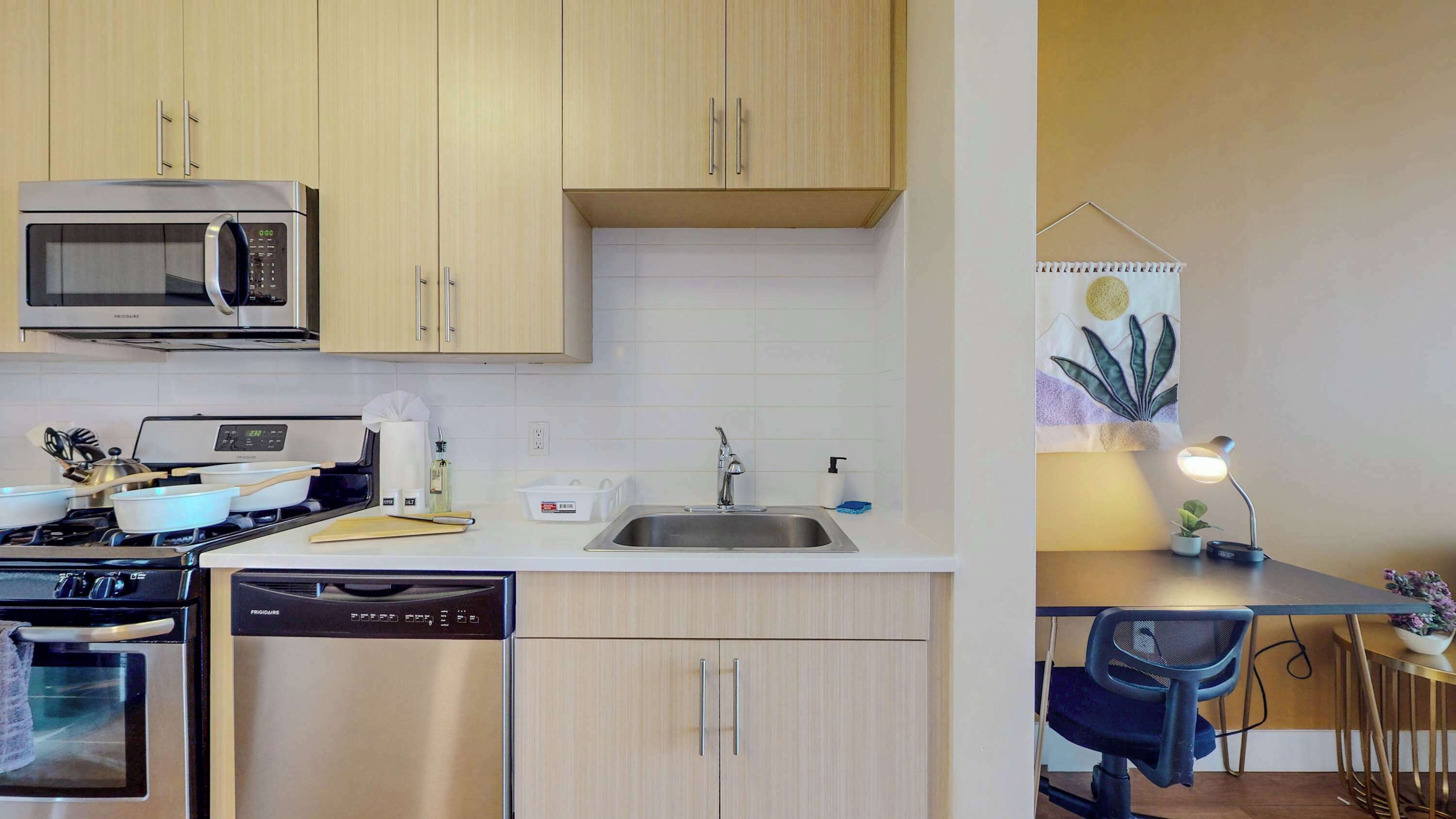A compact kitchen with a microwave, stove, and dishwasher. There is a sink with a faucet and a small table with a chair and a lamp.