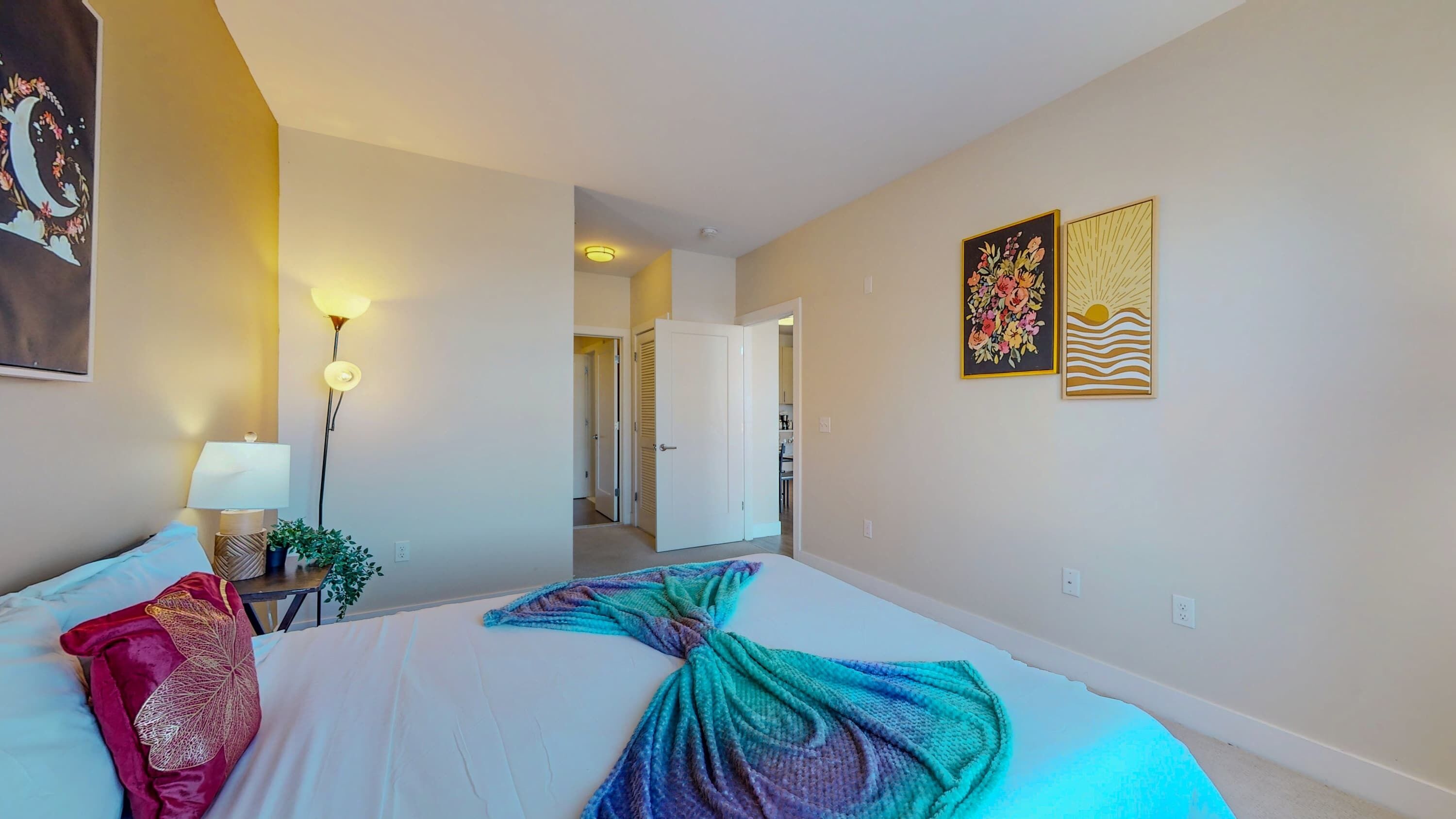 A hotel room with a large bed, a nightstand with a lamp, a potted plant, and two framed artworks on the wall.