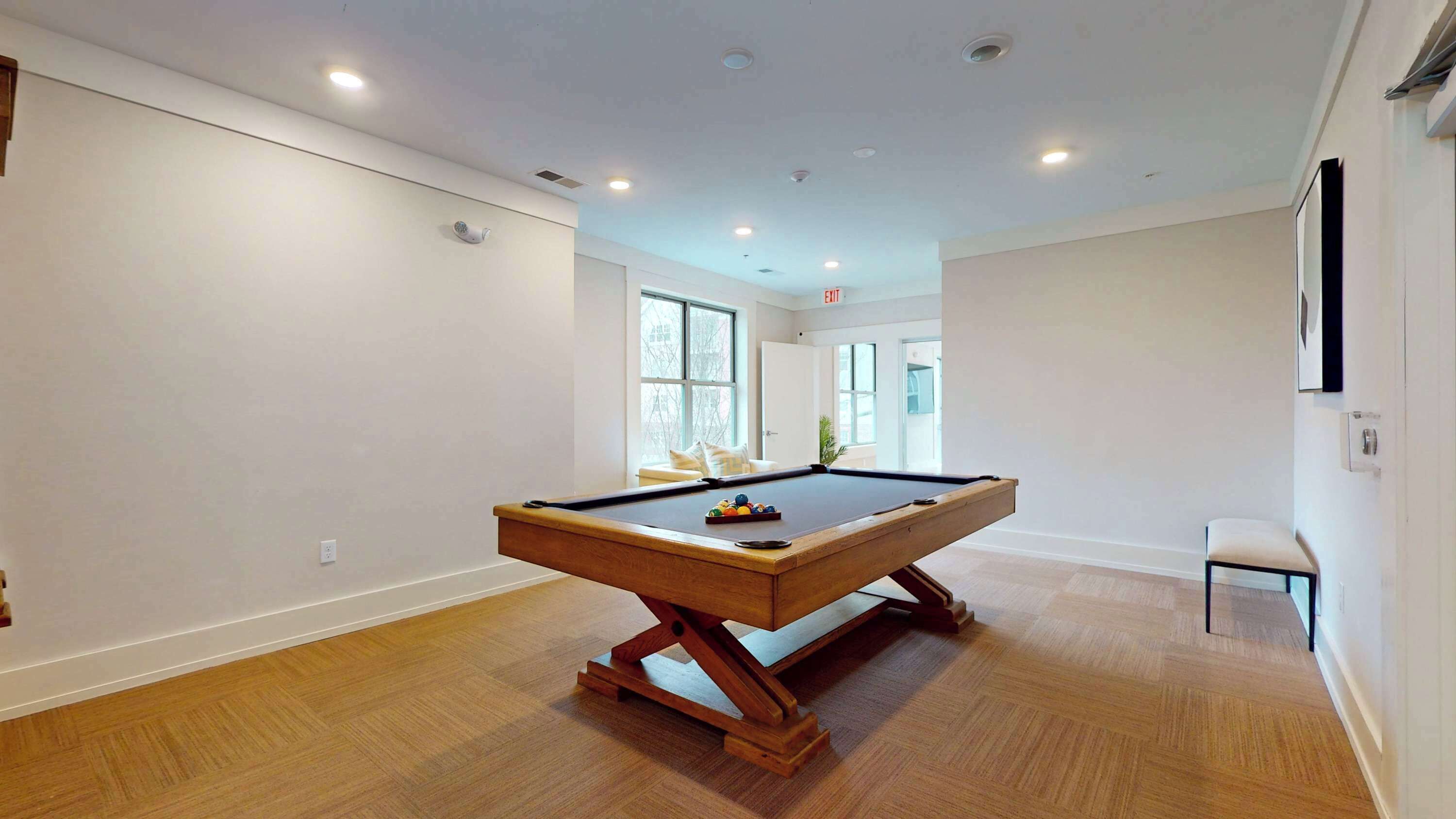 A spacious room with a pool table, wooden flooring, and large windows allowing natural light.