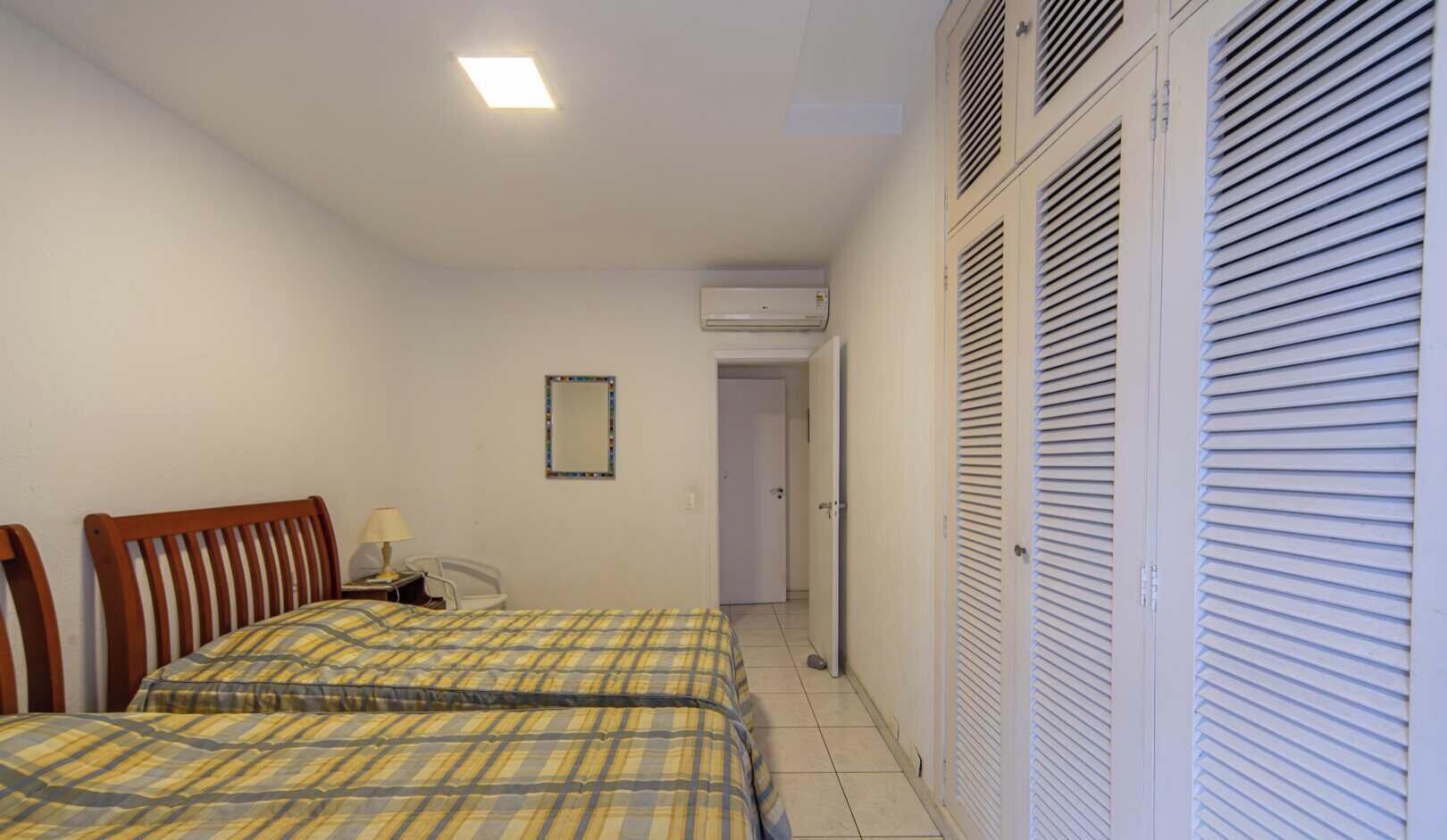 A bedroom with two beds, a bedside table with a lamp, a mirror, an air conditioning unit, and a door leading to another room.