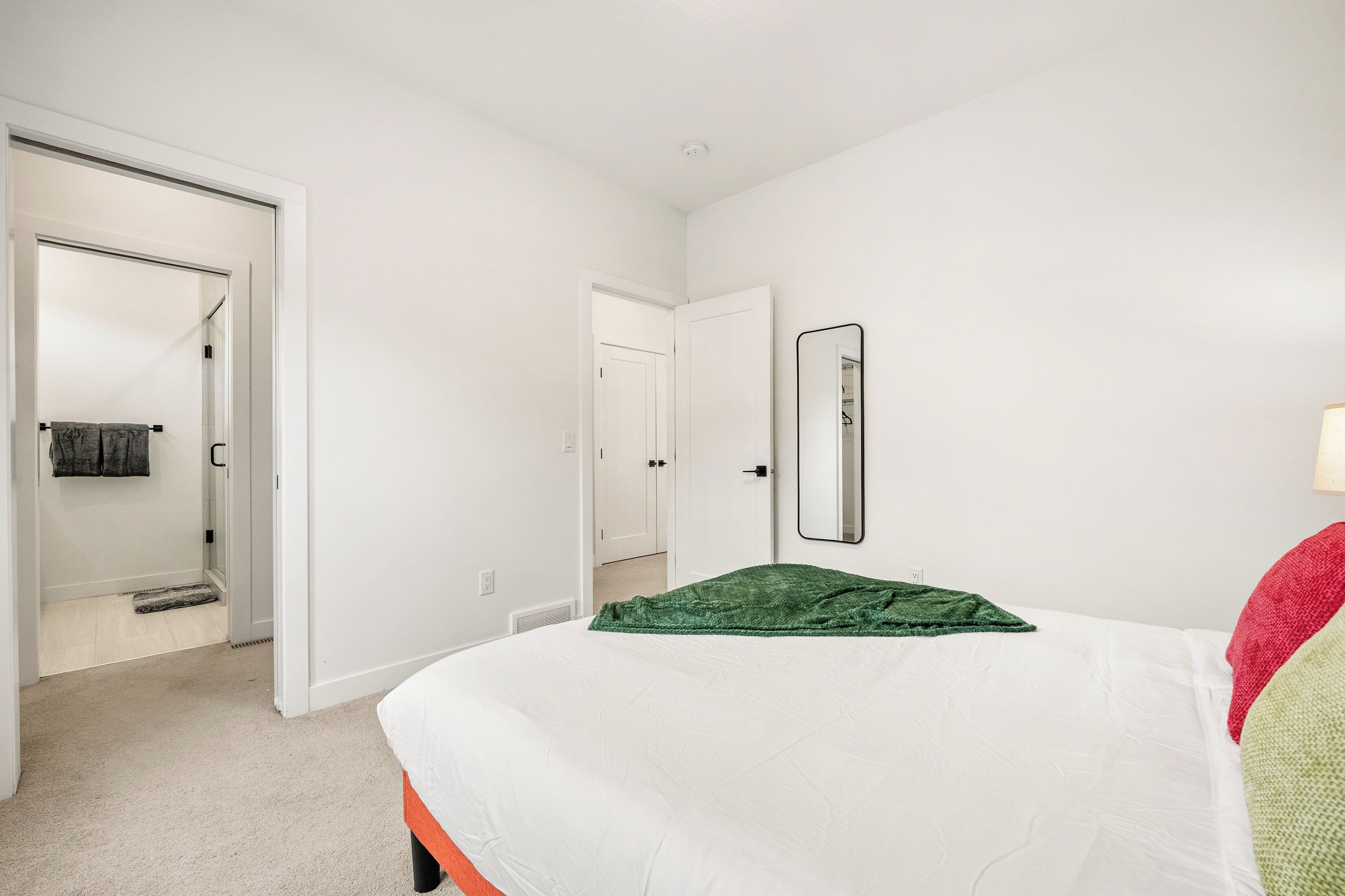 Une chambre à coucher avec un lit, un plaid vert, un mur blanc, un miroir et une porte donnant sur une autre pièce.