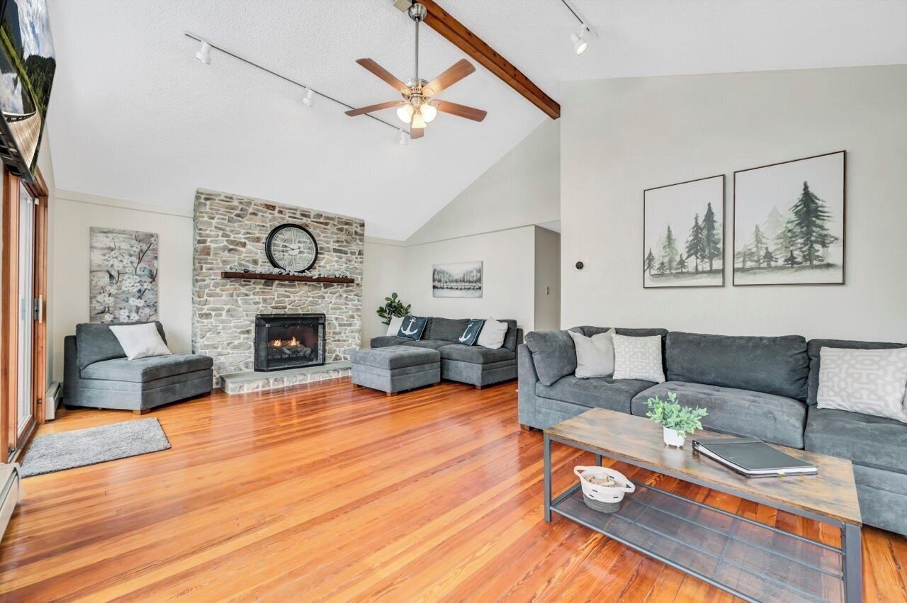 A spacious living room with a stone fireplace, wooden flooring, and a ceiling fan.