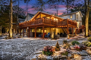 A two-story wooden cabin with a balcony, surrounded by trees and a snow-covered ground.