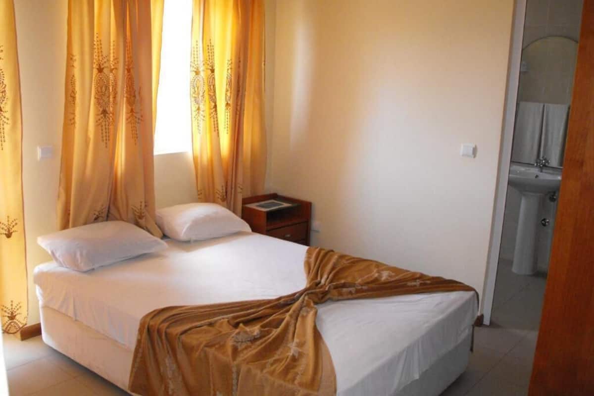 Un dormitorio con cama, cortinas, una mesita de noche y un baño visible a través de una puerta abierta.