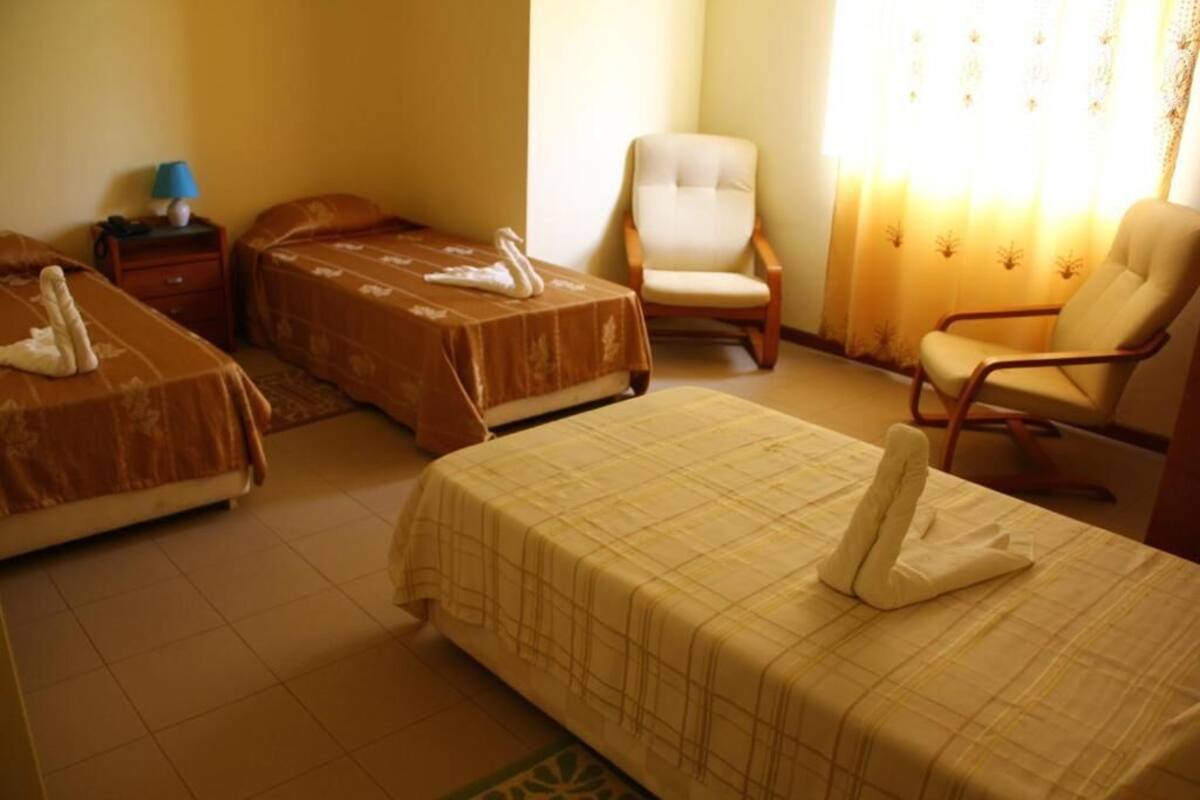 Habitación de hotel con dos camas, una silla, una mesita de noche con lámpara y una ventana con cortinas.
