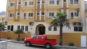 Un edificio de varios pisos con fachada amarilla y blanca, una camioneta roja estacionada al frente y una palmera a la derecha.