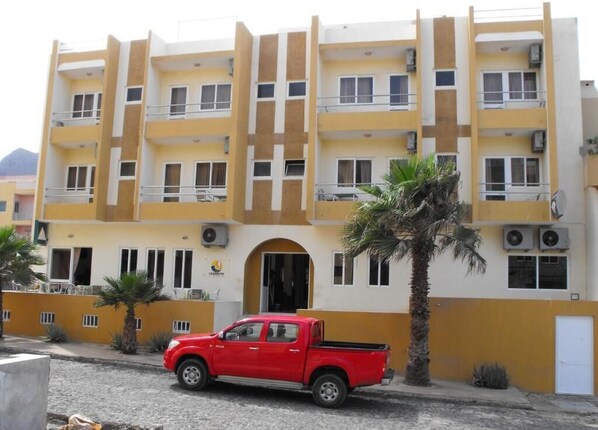 Ein mehrstöckiges Gebäude mit gelb-weißer Fassade, ein roter Pickup-Truck davor geparkt und eine Palme rechts.