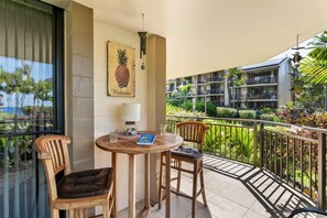 A balcony with a wooden table, two chairs, a pineapple artwork, and a view of a hotel complex. - Kona Makai 5-101: Ground Floor,1 Bedr/2 Bath! (Kailua-Kona)