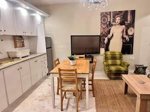 A modern kitchen with white cabinets, a dining area with a wooden table and chairs, a television, and a green armchair.