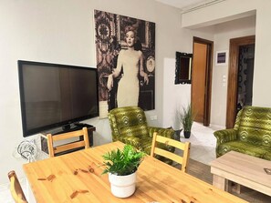 A living room with a flat-screen TV, a wooden dining table, two green patterned armchairs, and a large framed picture on the wall.