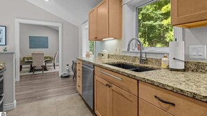 A modern kitchen with granite countertops, wooden cabinets, and a large window providing a view of greenery. - Relax & Recharge | Fire pit, Game Room, Near Torch Lake (Bellaire)