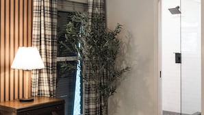 Un dormitorio con cama, mesita de noche, lámpara, planta en maceta, ventana con cortinas y puerta que da a un área de ducha.