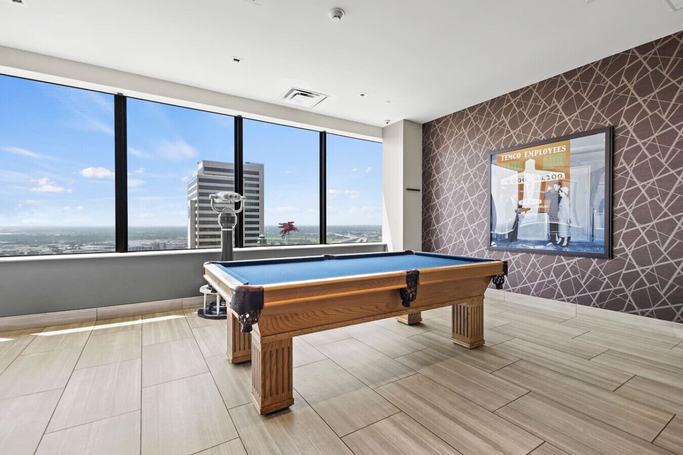 Une table de billard dans une pièce dotée de grandes fenêtres donnant sur la ville, une affiche accrochée au mur et un motif décoratif sur le mur.