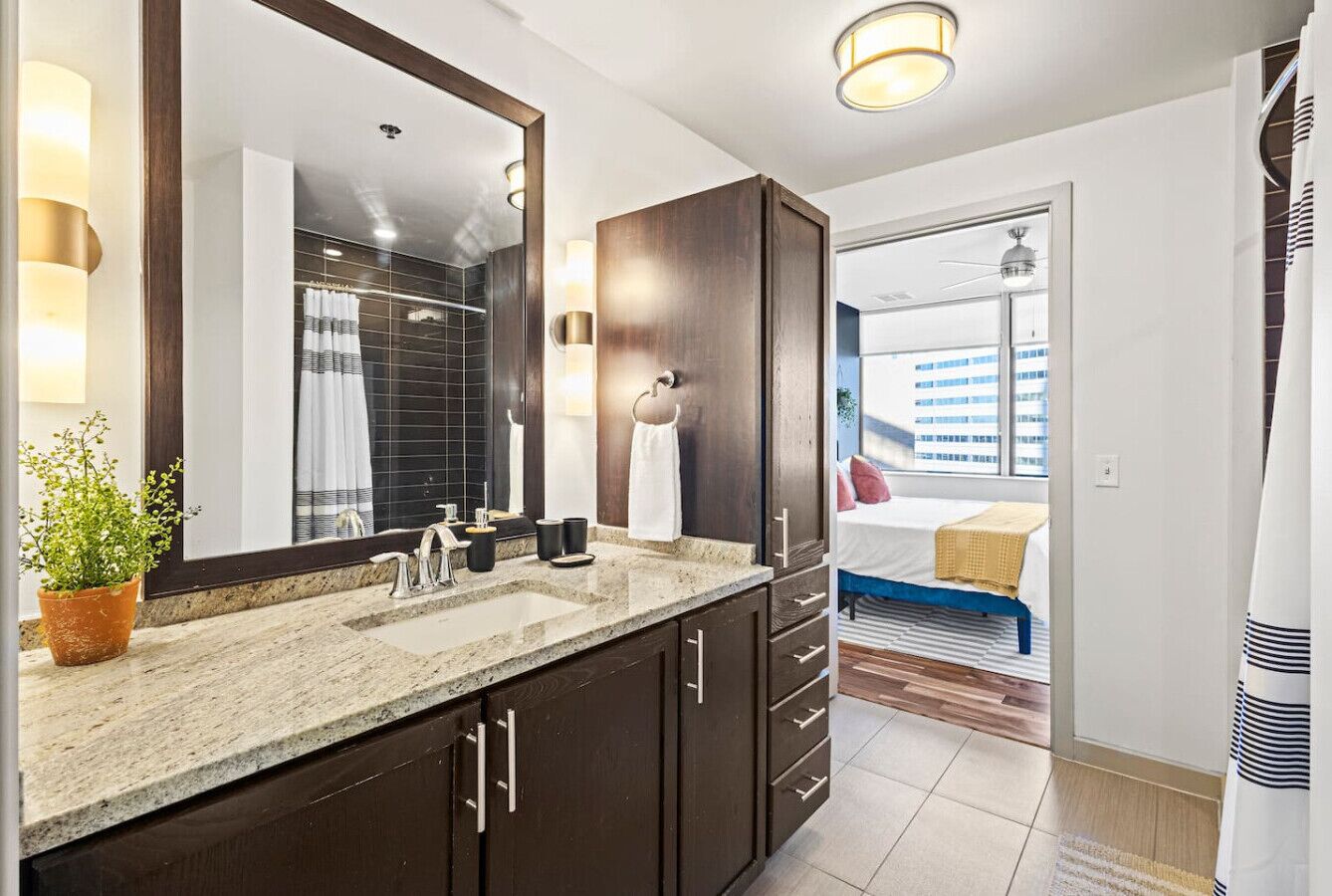 A modern bathroom with a large mirror, dark wooden cabinets, a granite countertop, and a potted plant.