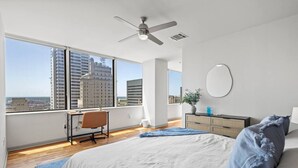 Una camera da letto moderna con un letto grande, una scrivania e una sedia. Offre vista sulla città attraverso ampie finestre, una ventola a soffitto e uno specchio rotondo sulla parete.