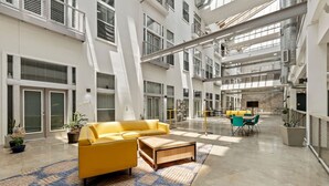 A spacious atrium with a yellow sofa, a coffee table, and a dining area with chairs and tables. - 2BR Modern Stay • Walk to AA Center• Free Parking (Dallas)