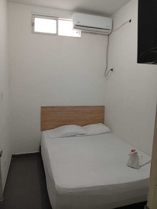 A small, single-bed room with a wooden headboard, a window, and an air conditioning unit mounted on the wall.