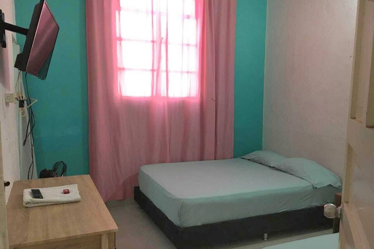 Una habitación pequeña y ordenada con una cama individual, una mesita de noche con un control remoto y una ventana con cortinas rosadas.
