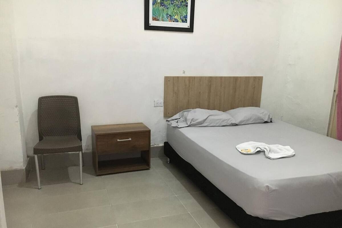 Una habitación pequeña y sencilla con una cama, una silla, una mesita de noche y un cuadro en la pared.