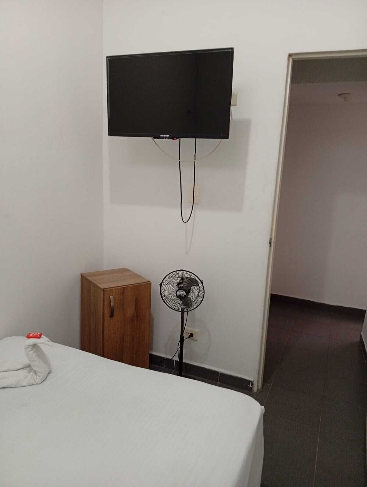 A room with a bed, a TV mounted on the wall, a wooden cabinet, and a standing fan.