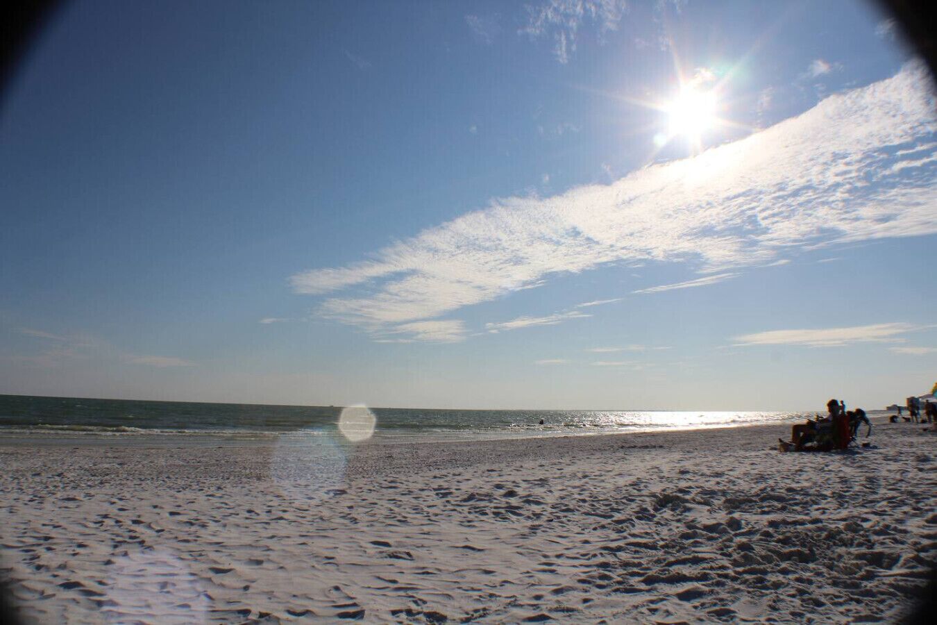 A beach with the sun shining, calm sea, and people relaxing on the sand.