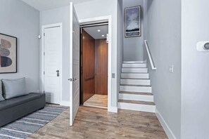 Ein Flur mit einem grauen Sofa, einem blauen Streifen Teppich, einer weißen Tür und einer Treppe, die zu einem Aufzug führt.