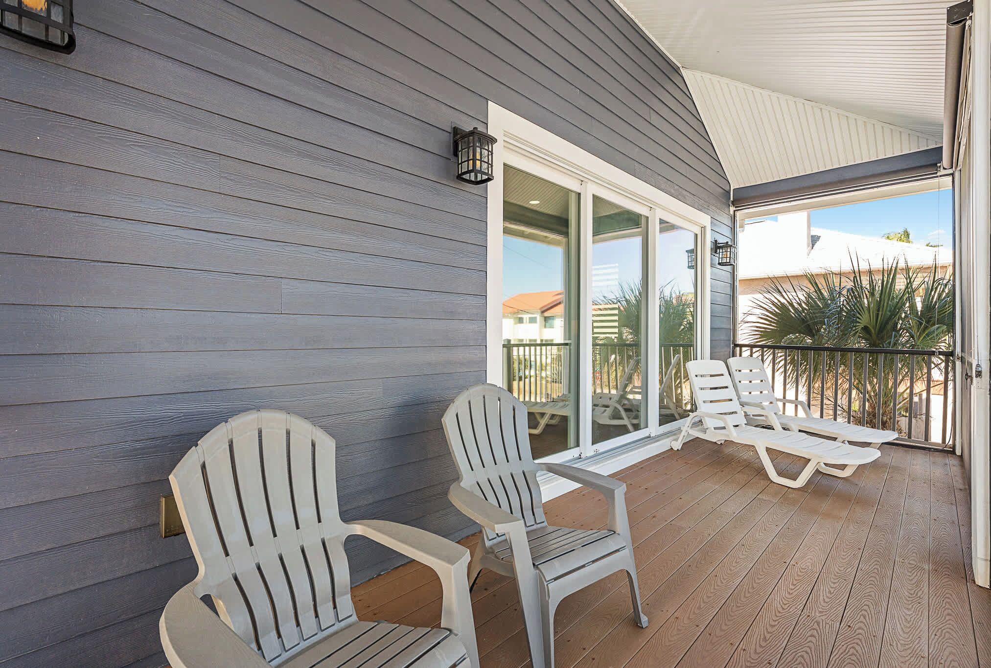 A covered patio with two Adirondack chairs and a lounge chair, a sliding glass door, and a view of a residential area.
