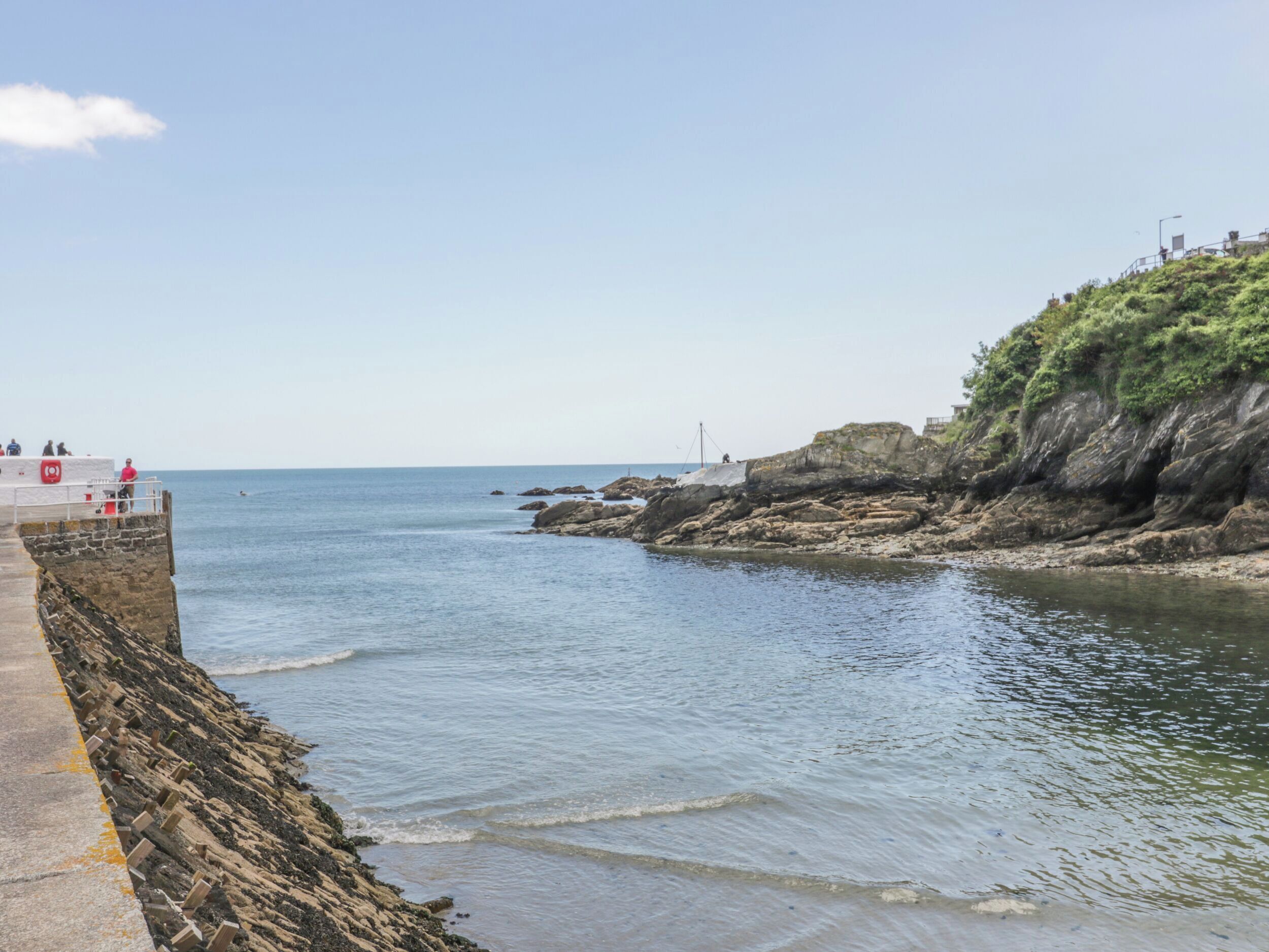 Un paysage côtier avec une rive rocheuse, une jetée s’avançant dans l’eau et un petit bâtiment sur la gauche, orné d’une bouée de sauvetage rouge.