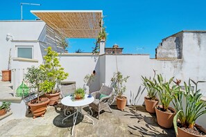 Een dakterras met plantenpotten, een ronde tafel, rieten stoelen en een pergola.