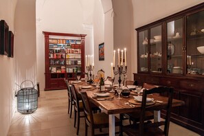 Une salle à manger avec une longue table en bois, dressée pour six personnes, entourée de chaises. On aperçoit une bibliothèque et un meuble avec des portes en verre à l’arrière-plan.