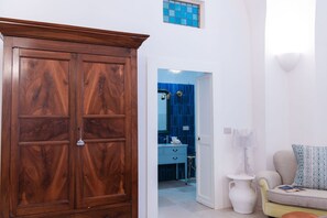 A room with a wooden wardrobe, a white door leading to a bathroom with a blue shower, a white pedestal table, and a sofa with patterned cushions.