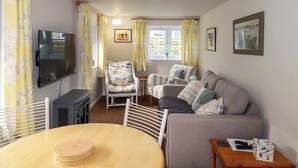 A cozy living room with a flat-screen TV, a dining table with a stack of coasters, a grey sofa, and a patterned armchair. - The Penthouse (Looe)