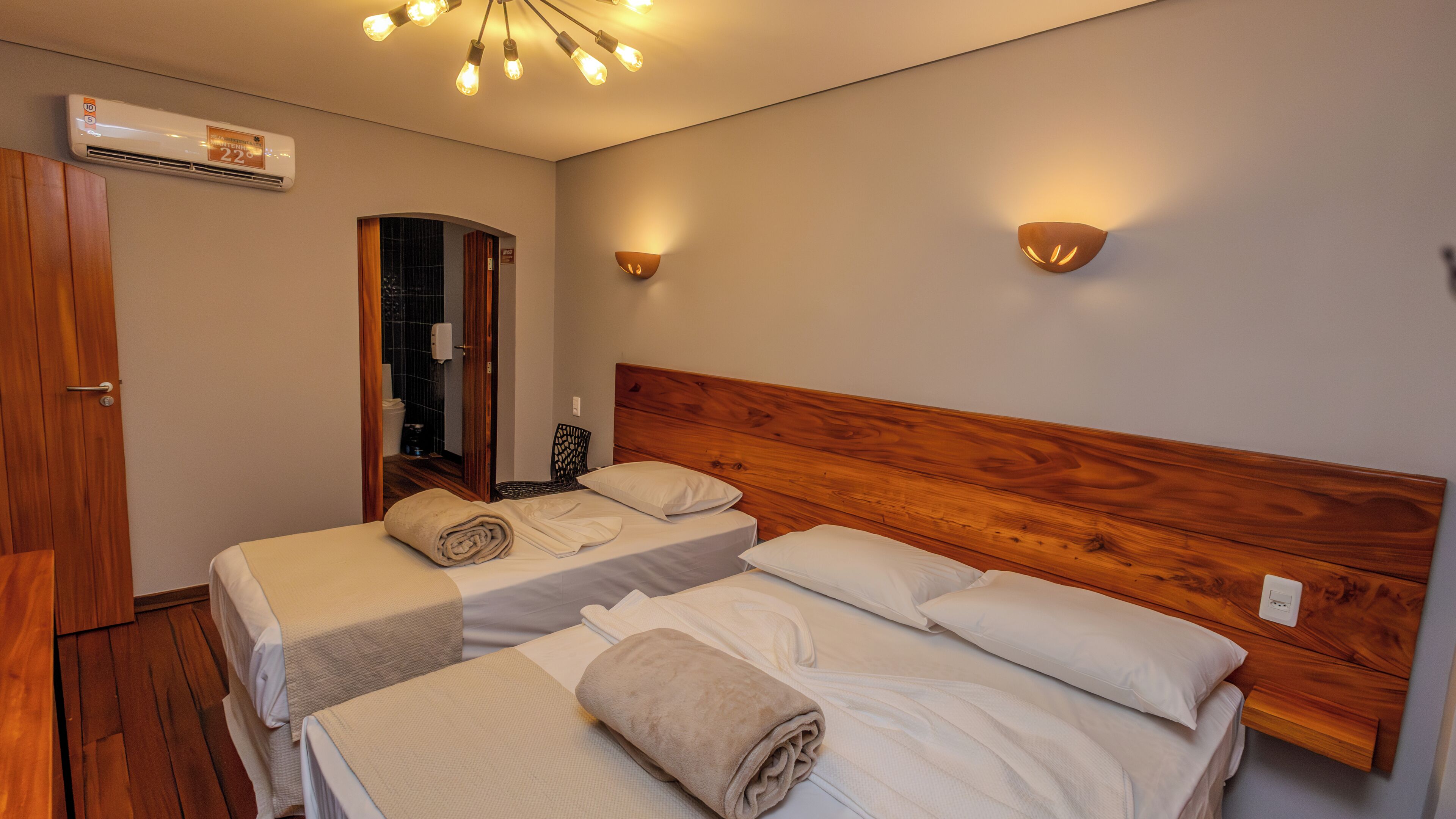 A hotel room with two beds, a wooden headboard, a mirror, a wall-mounted air conditioner, and a ceiling light fixture.