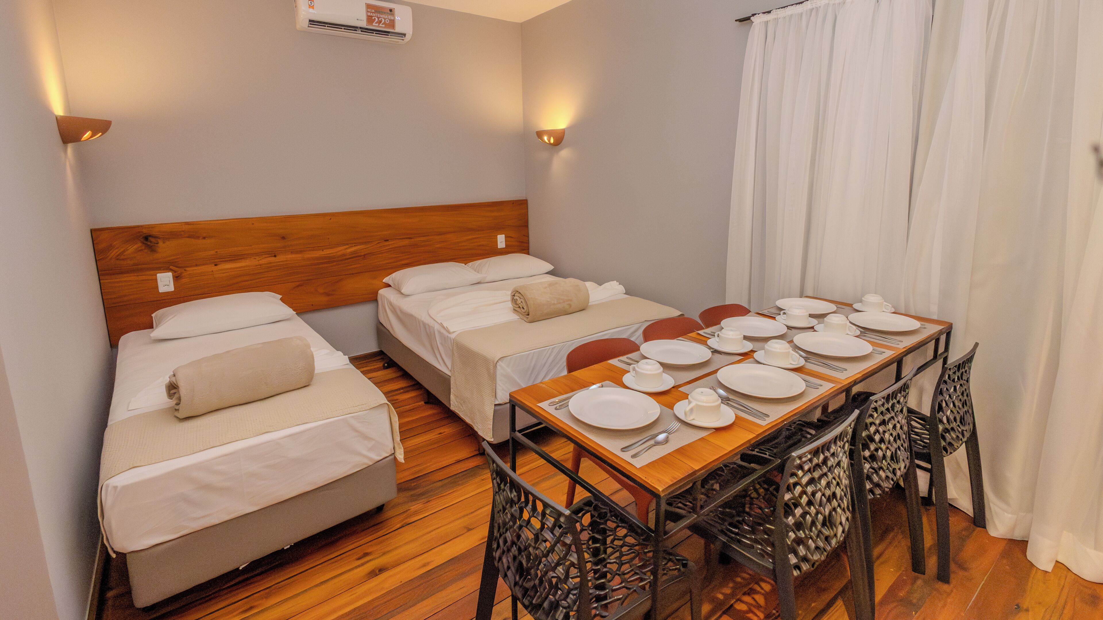 A hotel room with two single beds, a wooden headboard, a dining table set for four, and a window with white curtains.