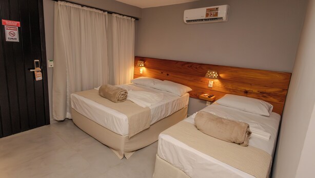 A hotel room with two beds, a wooden headboard, bedside lamps, an air conditioning unit, and a curtain. - Ap Passarela (Porto Seguro)