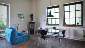 Een gezellige kamer met een houten vloer, een blauwe relaxfauteuil, een eettafel met stoelen, een open haard en grote ramen met uitzicht op groen.