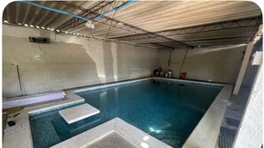An indoor swimming pool with a tiled floor and walls, a diving board, and a ceiling with exposed beams and lighting.