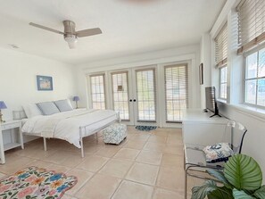 A bedroom with a bed, bedside tables, a desk, a chair, a ceiling fan, and large windows with blinds.
