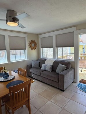 A living room with a gray sofa, wooden dining table, and chairs. There are large windows with blinds, a ceiling fan, and a decorative sunburst wall piece.