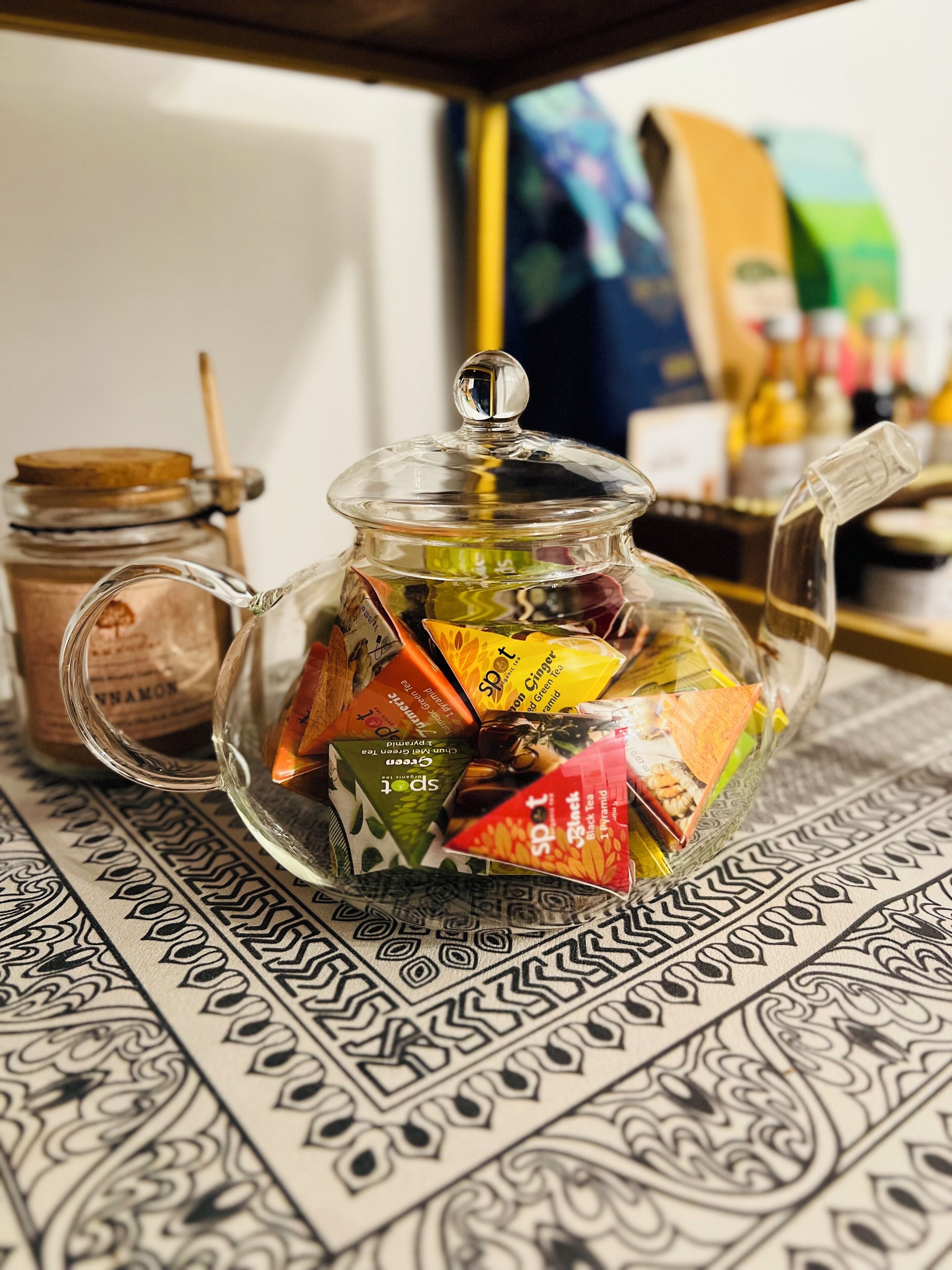 A glass teapot filled with tea bags on a patterned tablecloth.