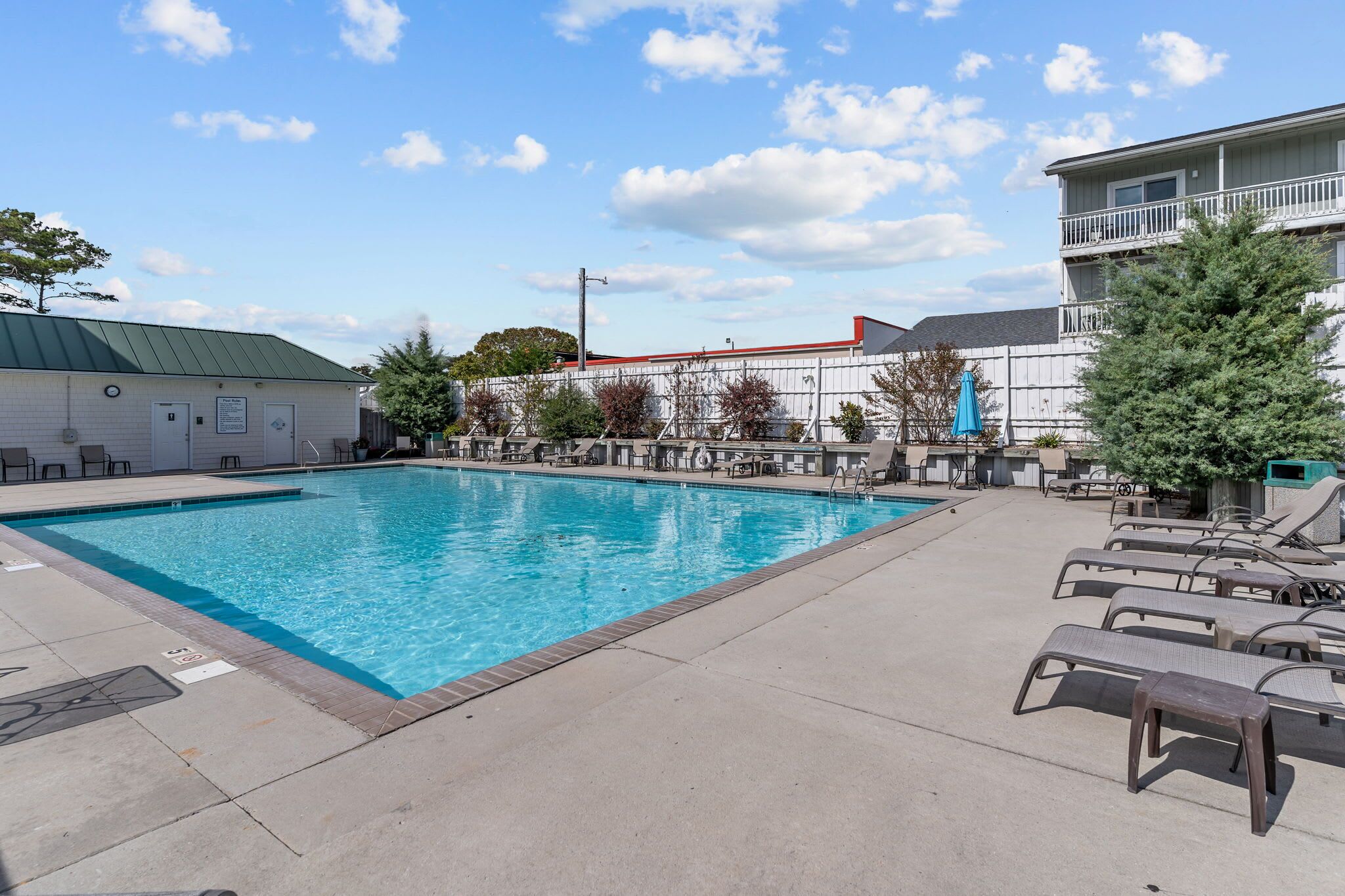 Une piscine rectangulaire, bordée d’une terrasse carrelée, entourée de chaises longues et d’une clôture en bois.