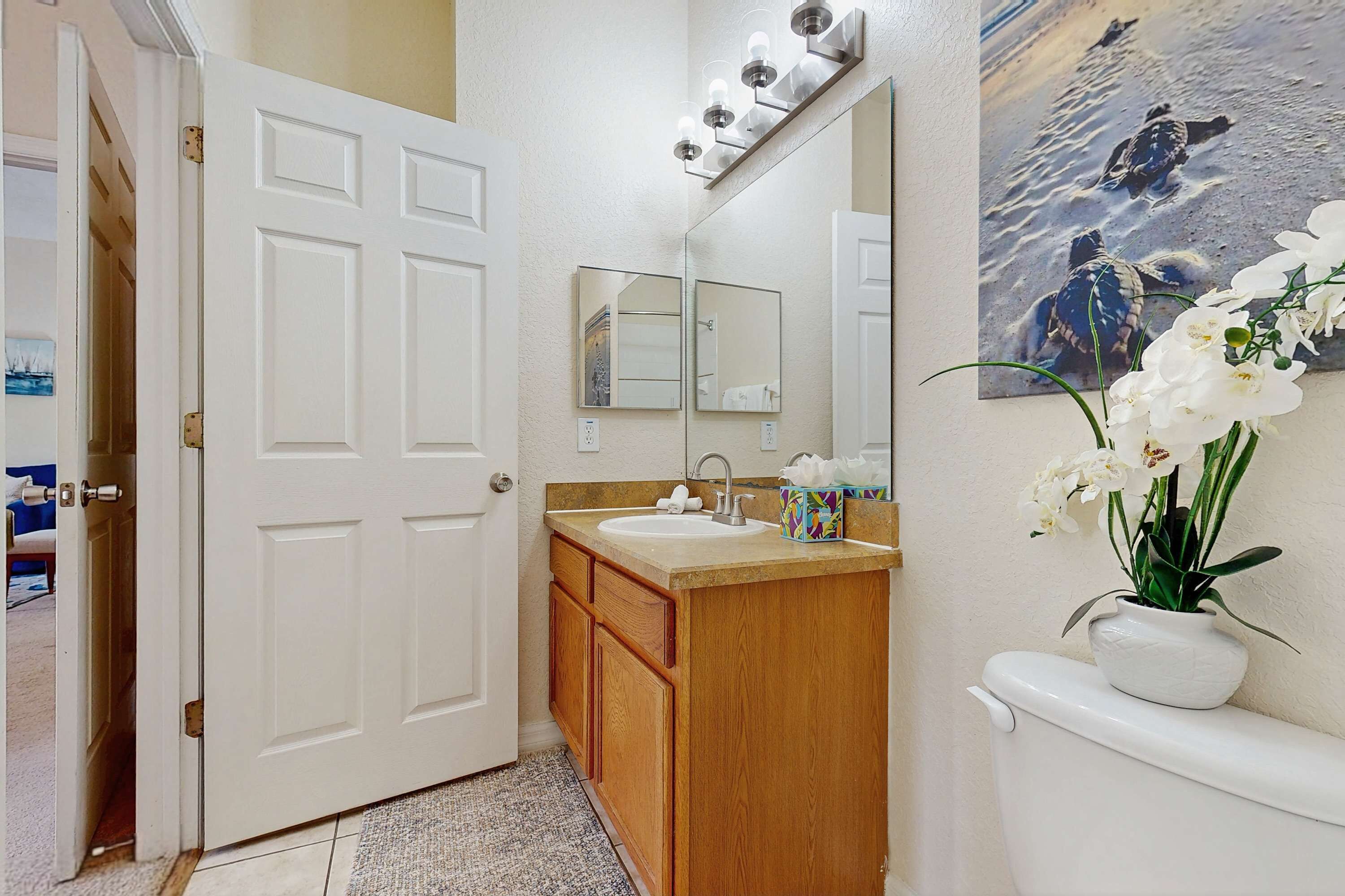 A bathroom with a white door, wooden cabinets, a sink, a mirror, a toilet, and a wall-mounted light fixture.